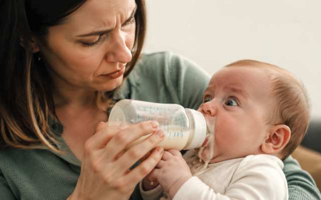 A concerned mother holding a baby bottle while feeding her infant who is spilling milk.
