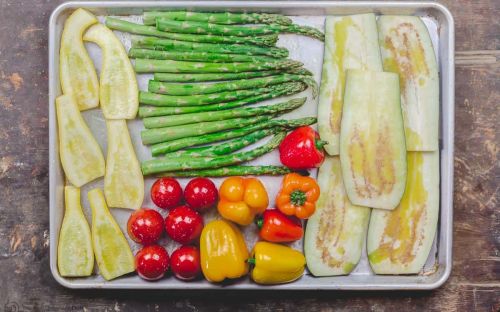 a baking tray is covered with long slices of yellow squash and eggplant stalks of asparagus and whole bell peppers