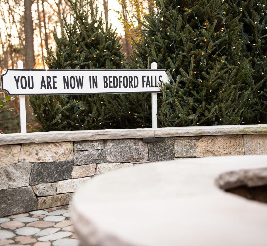 a knee wall hosts a sign for a Bedford Falls town and stand in front of lit Christmas trees