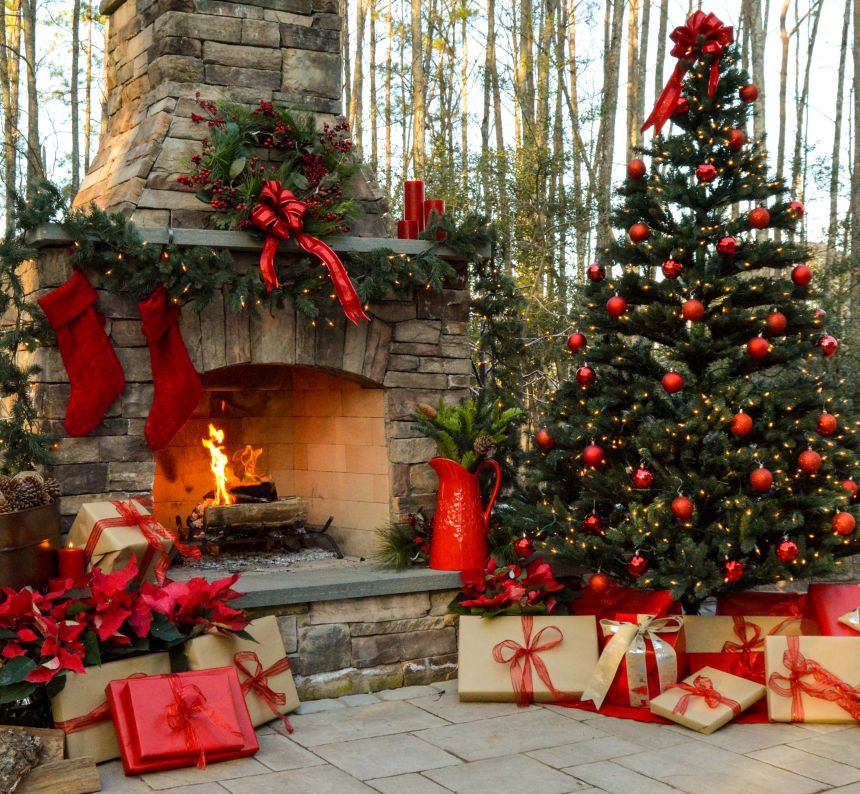 Large Christmas tree sits beside the large stone fireplace with Christmas gifts littering the patio all decorated with red ornaments holly berries green garland and wreaths