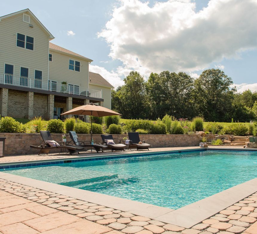 Large pool is surrounded by a paver patio with 4 lounge chairs nearby under an umbrella as an oasis in this backyard