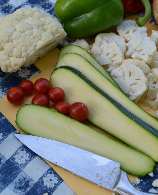 a dirty chef's knife sits on a yellow cutting board and beside long slices of zucchini, some cherry tomatoes and slices of cauliflower all ready for the grill