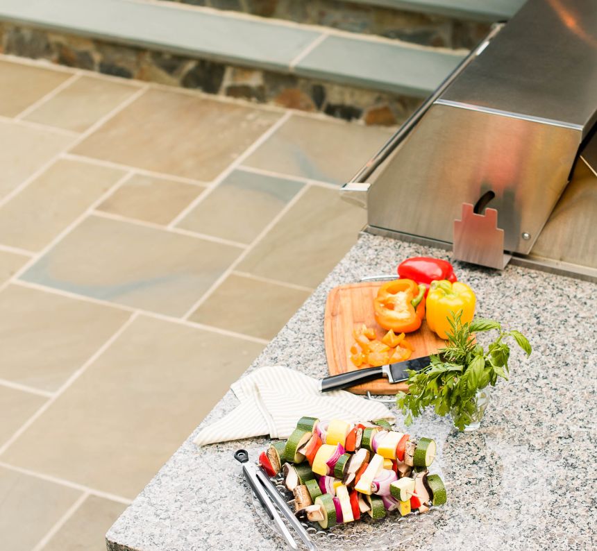 backyard stone paver patio with grill island kitchen and granite countertop has veggies ready to be grilled resting on it.