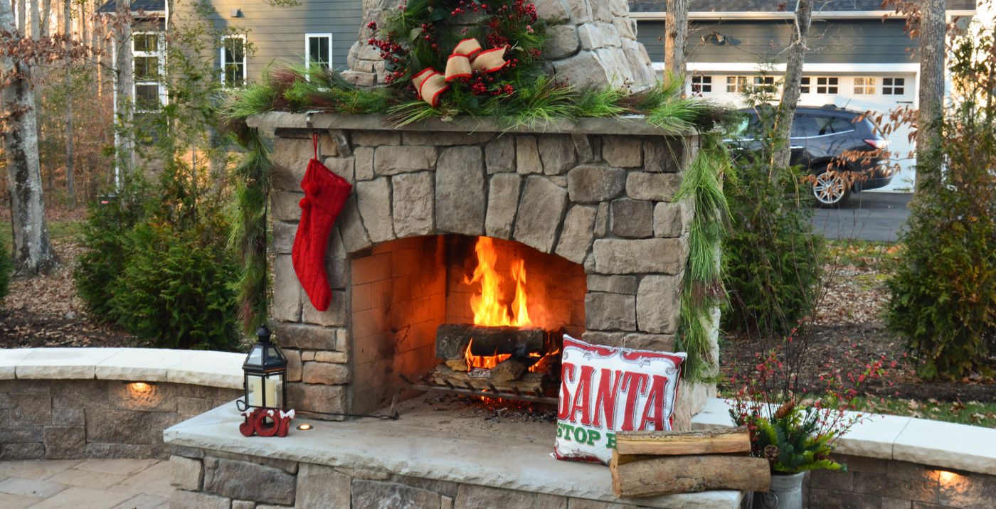 this outdoor stone fireplace hosts a roaring fire in mid day and is decorated with firewood, a Santa pillow, a stocking hanging from the mantle and the garland drapes over the mantle