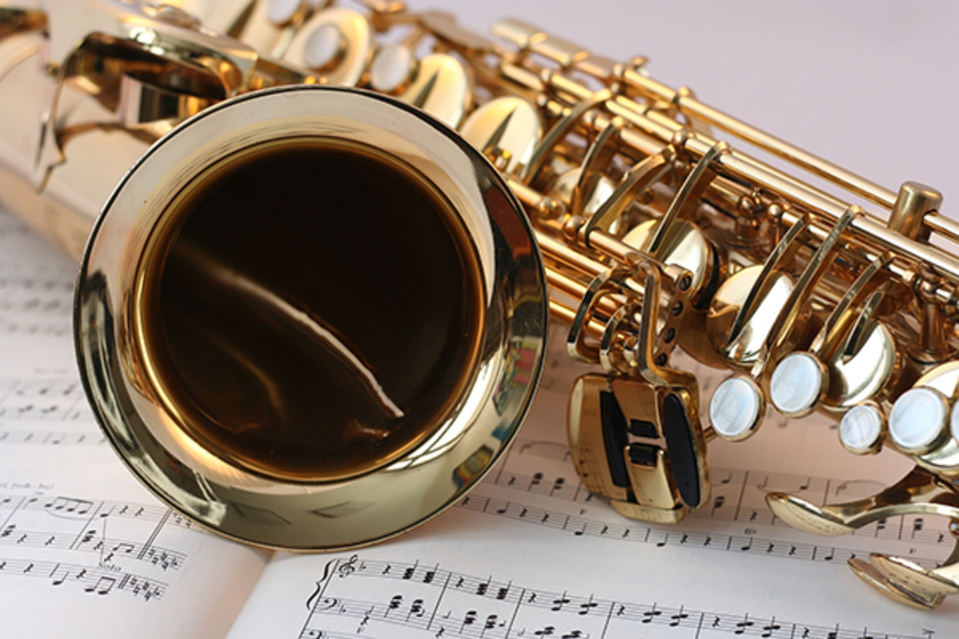 A saxophone lays on an open book of music. There is an off-white background.