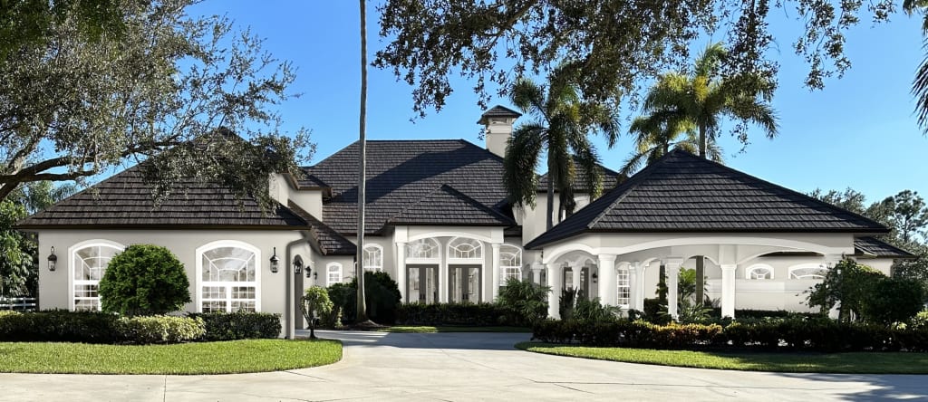 Coastal stucco home with arched windows, brown front doors, a carport, and a black roof.