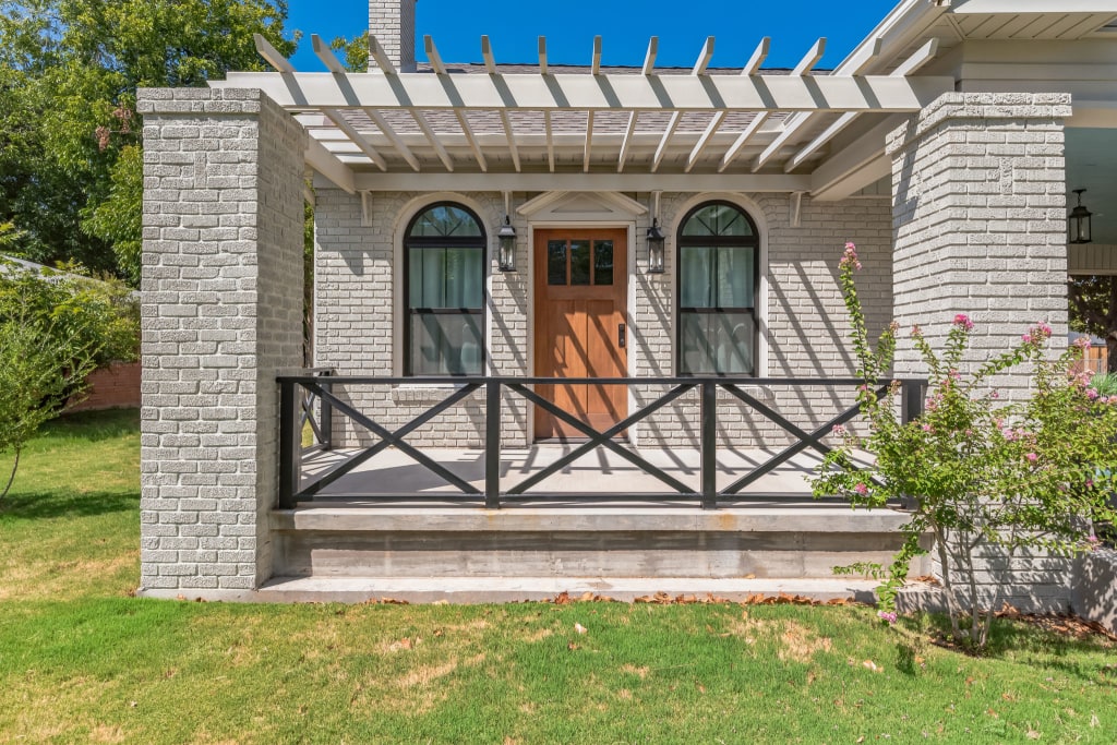The front porch of a home with a pergola, black railing, and wooden door.