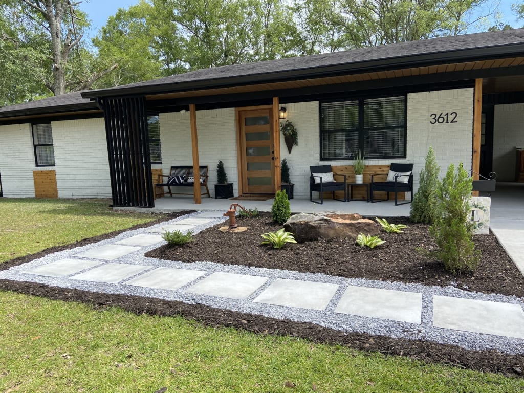 Single-story, white painted brick home with with a black roof and wooden accents, along with a stone walkway.