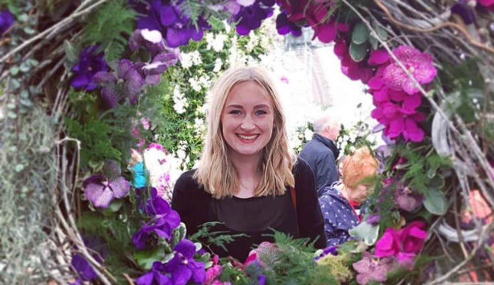 Young, blonde woman smiling at camera through a large flower wreath. 