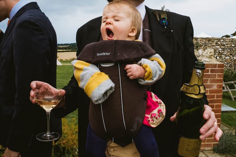 dad carries his baby at a wedding as he has a glass of champagne