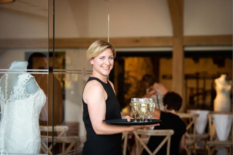 Woman in black dress holding tray with glasses of champagne and smiling at the camera.