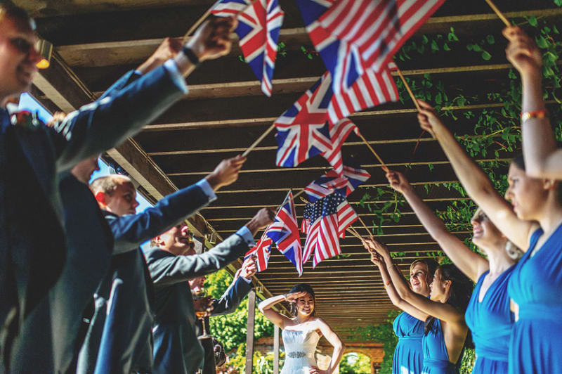 Bridebook.co.uk- bride standing under american and british flags held by wedding team