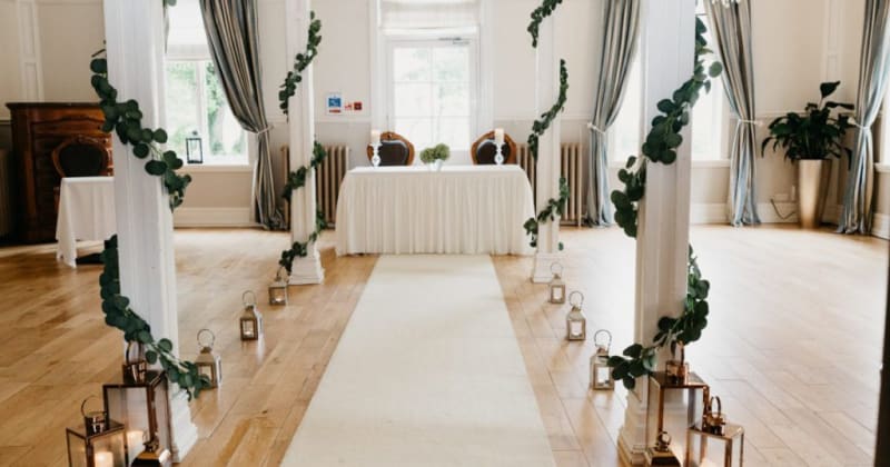 Image of ceremony room. Pillars down aisle with ivy wrapped around, a white aisle and table at the end with two chairs
