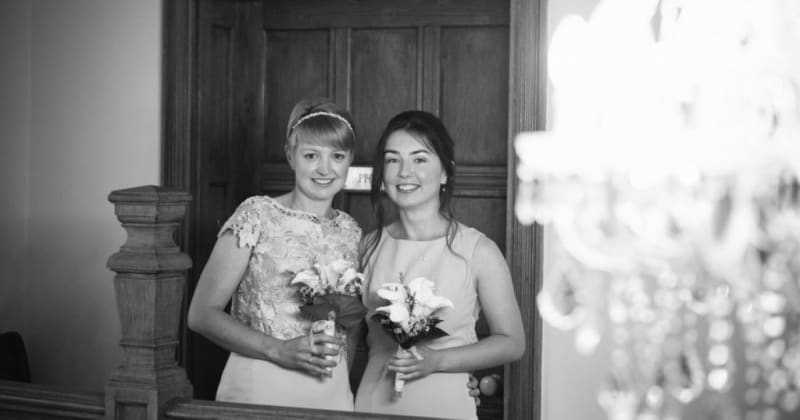 Black and white picture of brides smiling, holding bouquets inside house, in front of a closed wooden door
