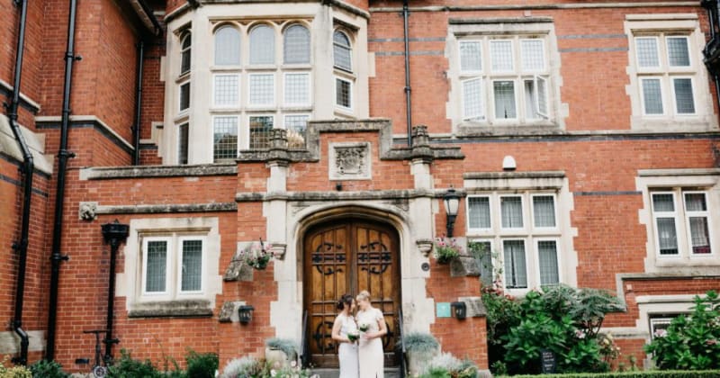 Brides outside large old building, smiling at each other with their bouquets in hand