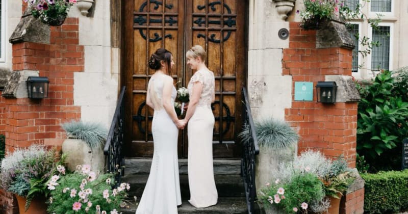 Brides outside wooden doors of Berwick Lodge, looking at each other smiling, holding hands. 