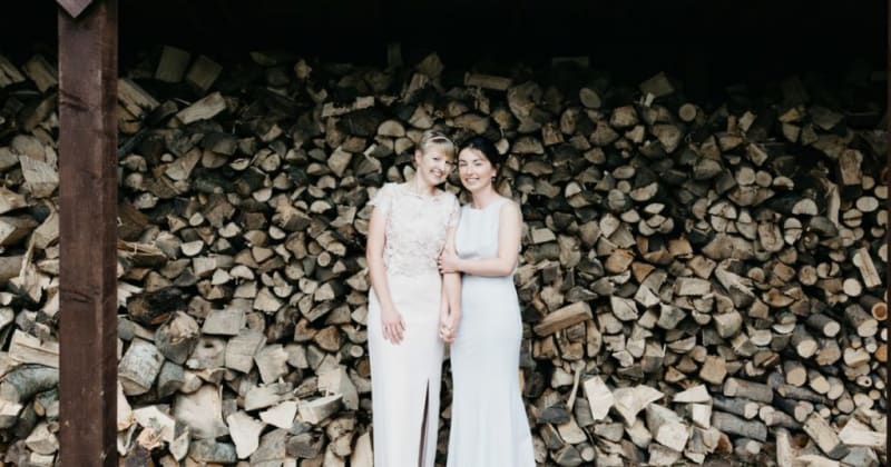 Brides smiling, holding hands in front of large stack of logs 