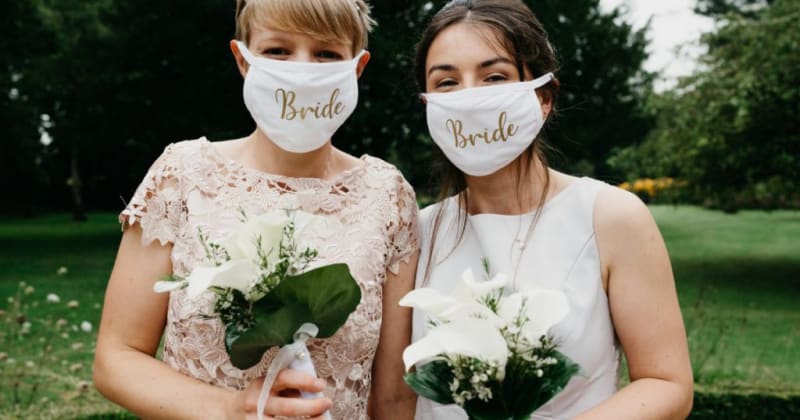 Brides smiling for camera in garden holding bouquets. Brides wear face masks with 'Bride' written on in gold