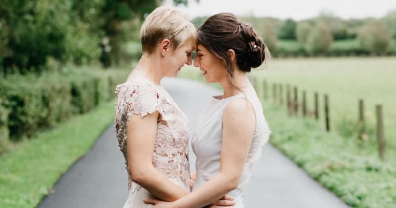 Image focused on brides, holding each other's hips and touching foreheads, with a long country road going off in the distance