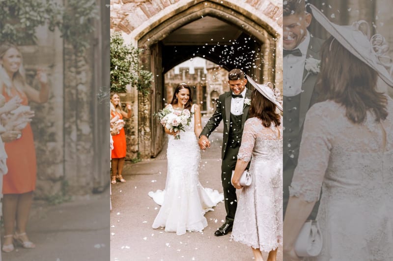 The bride and groom step outside smiling and holding hands as guests throw petals at them.