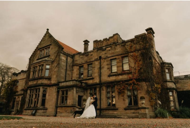 The bride and groom kiss in front of the building, which is made of stone. The colors are very autumnal.