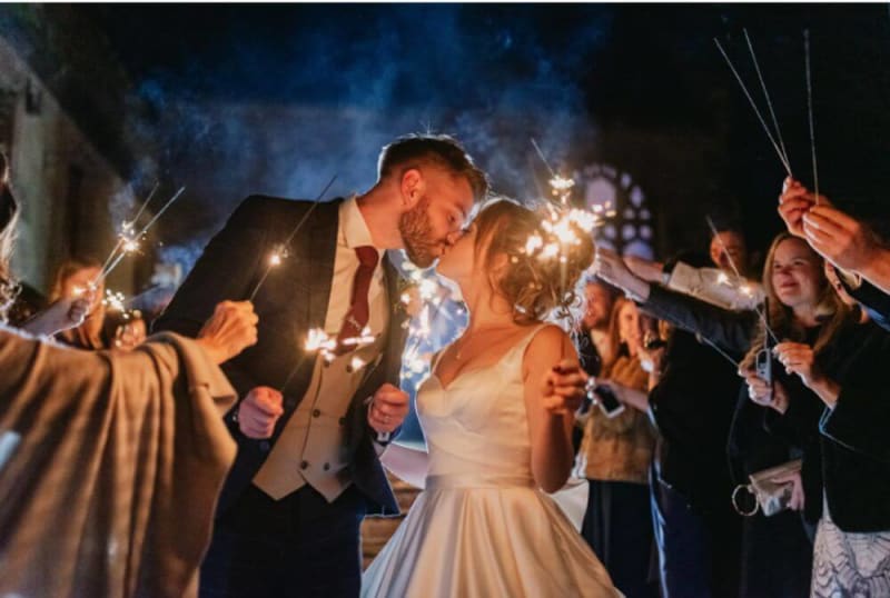 The bride and groom kiss outside at night as the guests hold candles.