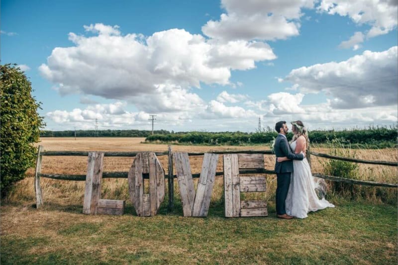 The couple embrace next to a big wooden sign that spells "LOVE".