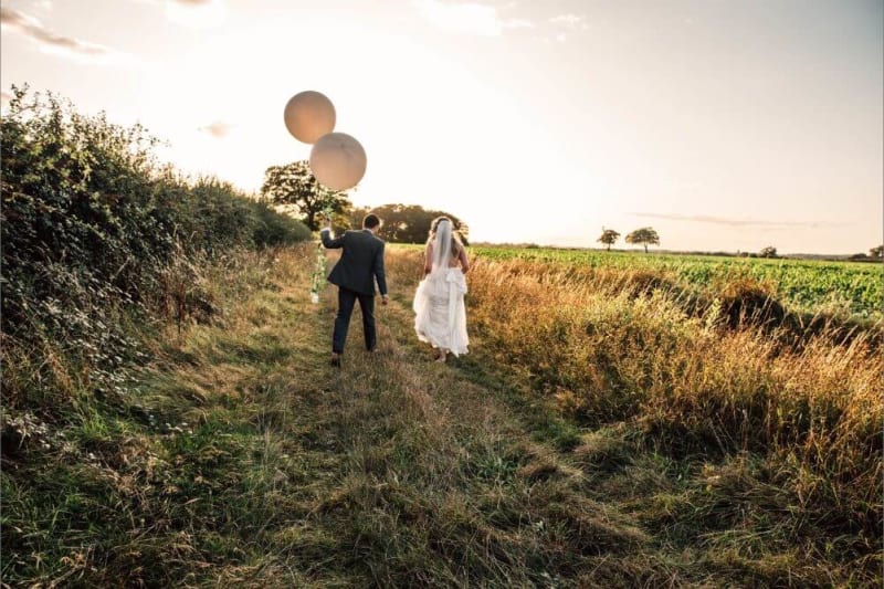 The bride and groom running outside as the groom holds two big white ballons.