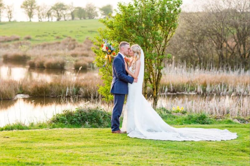 The couple kiss outside beside a lake.