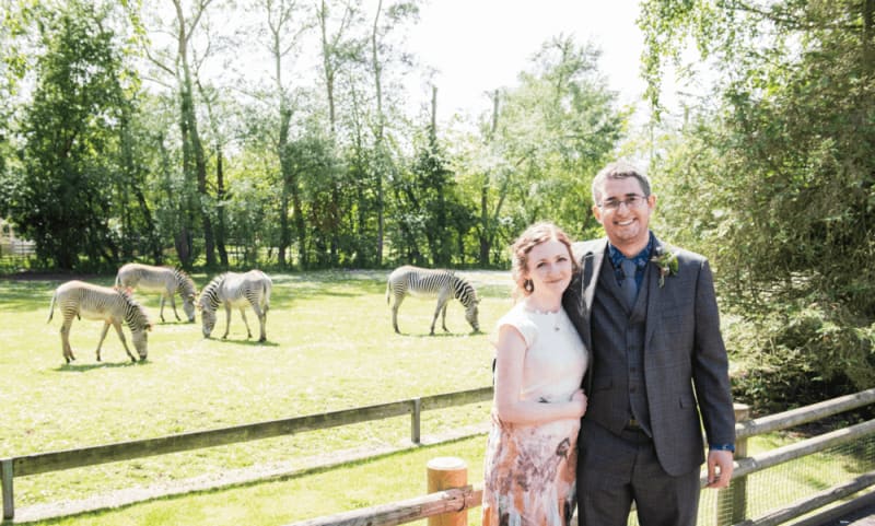 A couple stands and smiles for a picture with zebras in the background.