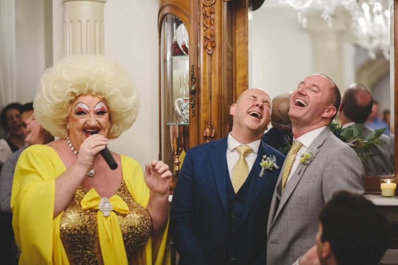 Man in drag with microphone next to two laughing men. 