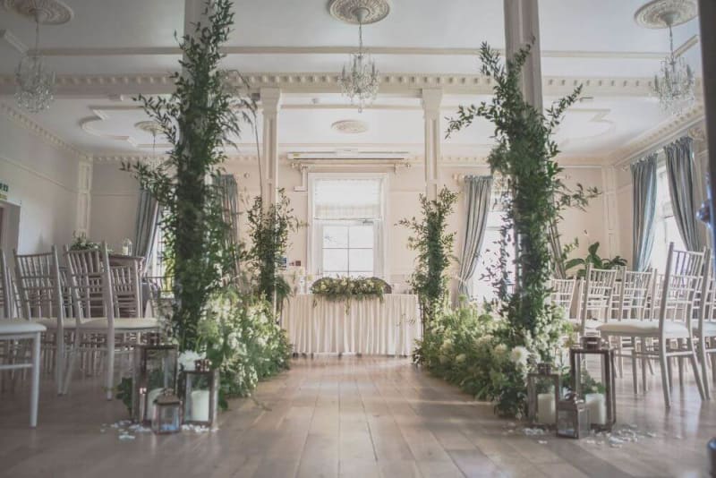 A hall with columns and chandeliers and chairs set up and decorated with flowers for a wedding ceremony. 