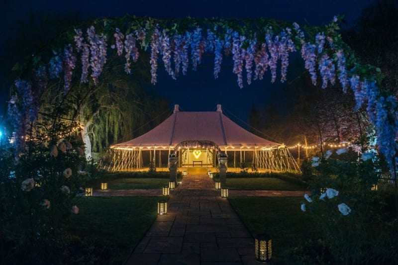 A white tent at night appears surrounded by lights. Purple flowers cover the frame.