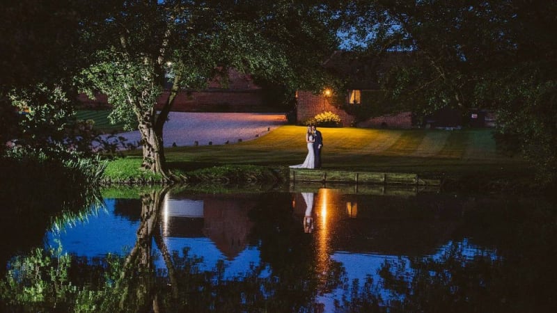 In the evening, the Bride and Groom stand beside a lake.