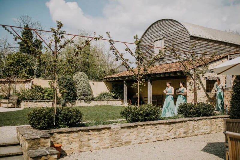 A procession of bridesmaids in blue dresses leaves a stone house.