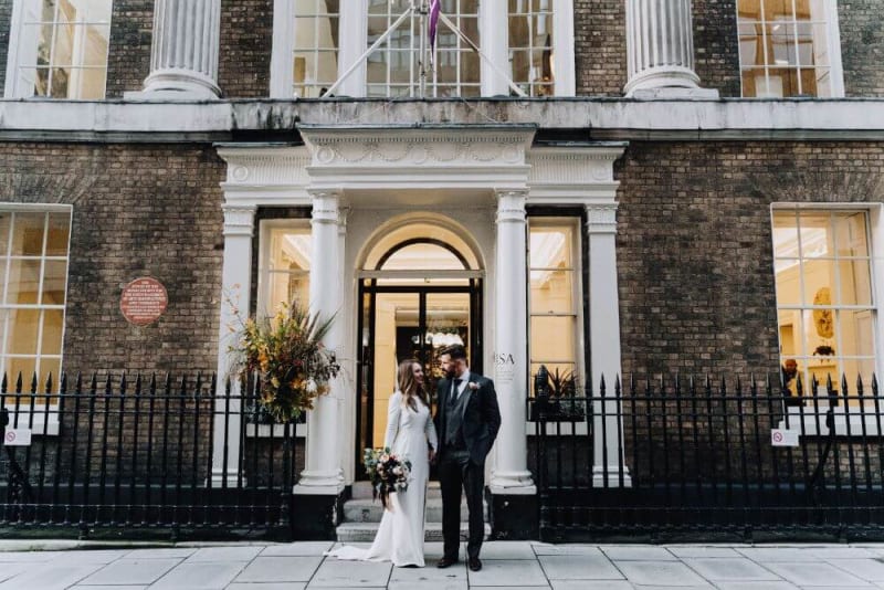 Bride and groom smiling at each other in front of the entrance to the venue. Bride is holding a bouqet.