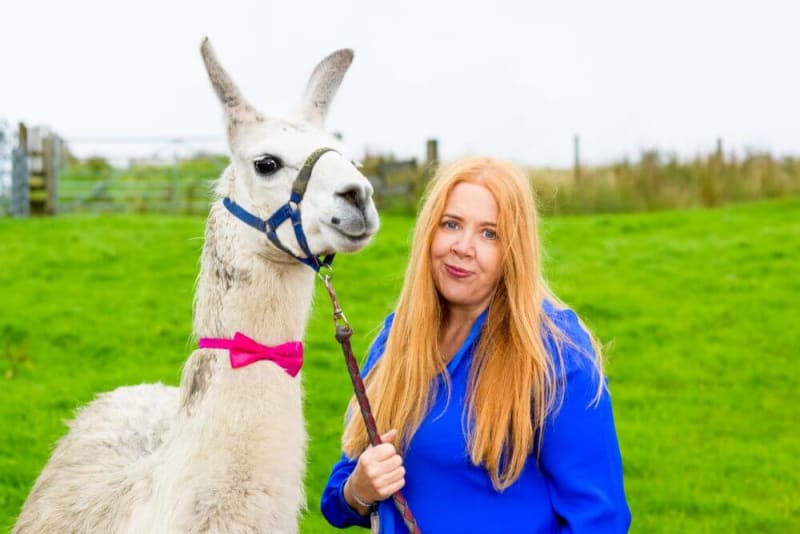 Woman smiling into camera next to a llama in a bow tie. 