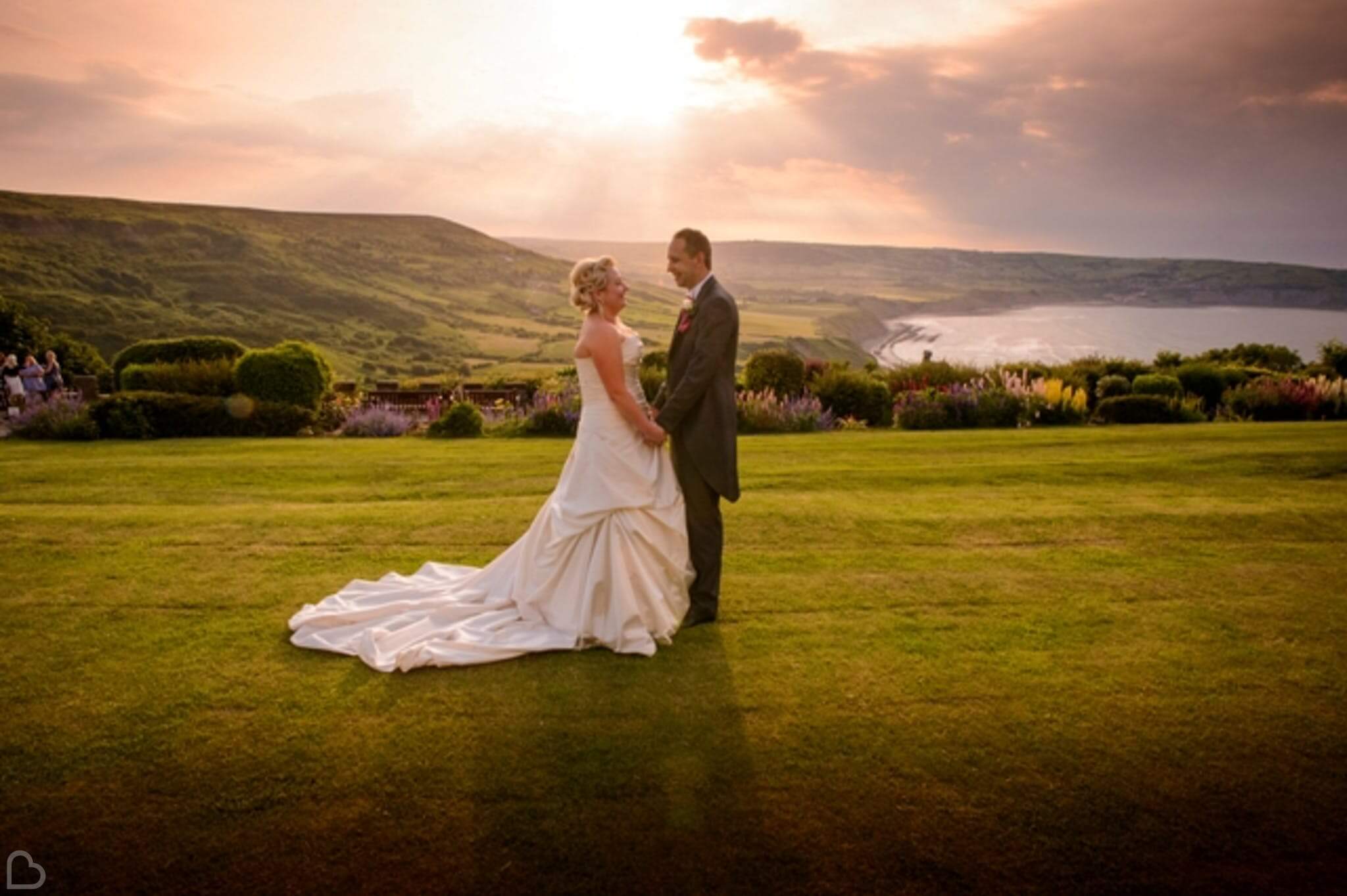 newlyweds hold hands at raven hall country house hotel, the sun shines over a lake in the background. 