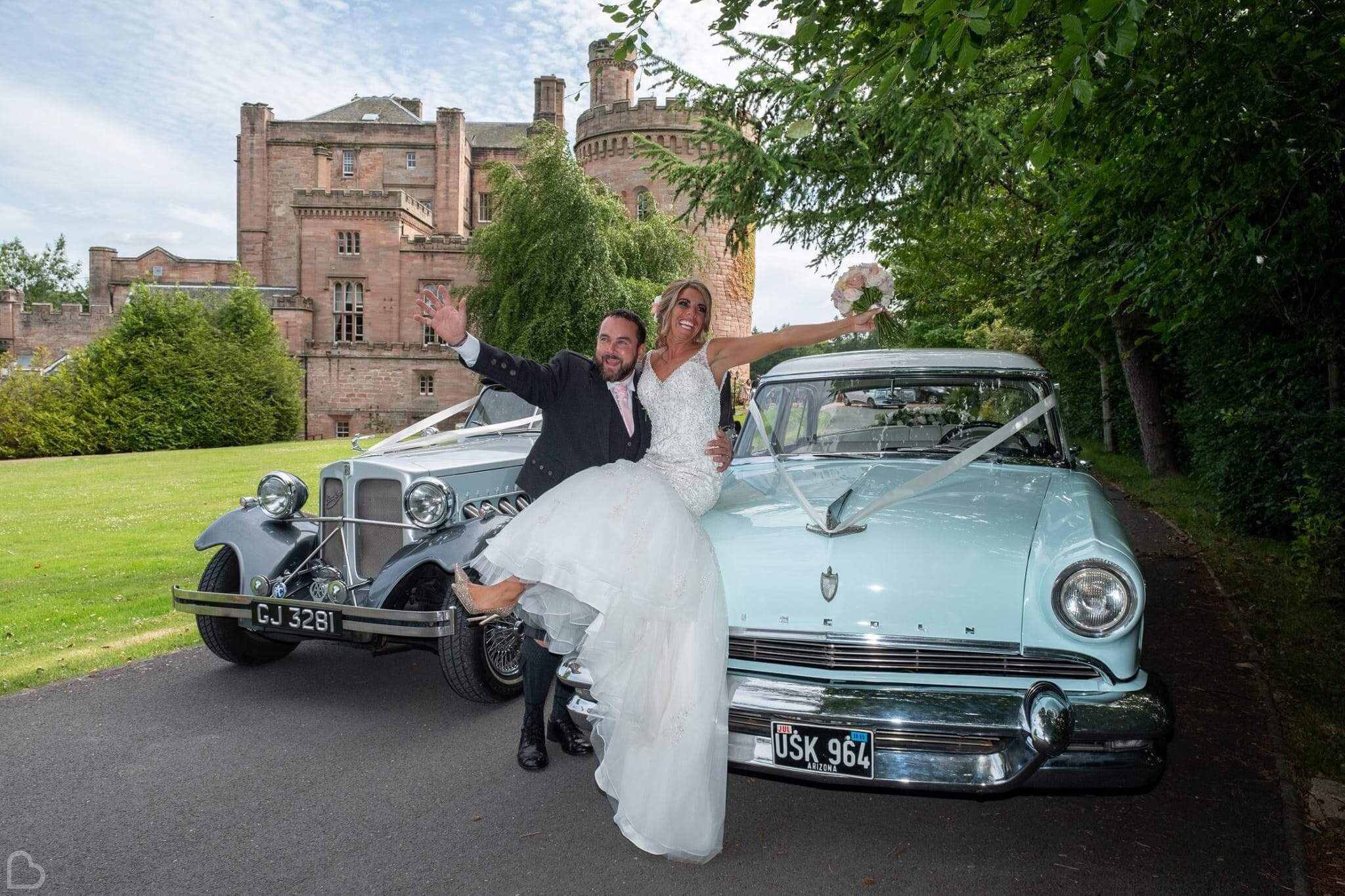 Newlyweds pose on their antique cars, in front of Dalhousie Castle.