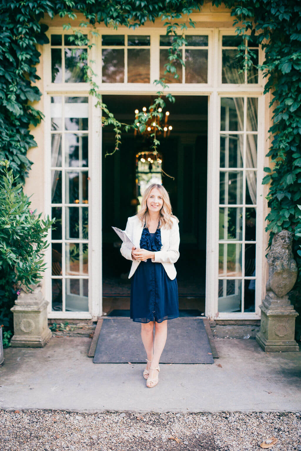 Smartly dressed woman standing in front of tall glass doors with a clipboard and smiling at camera. 