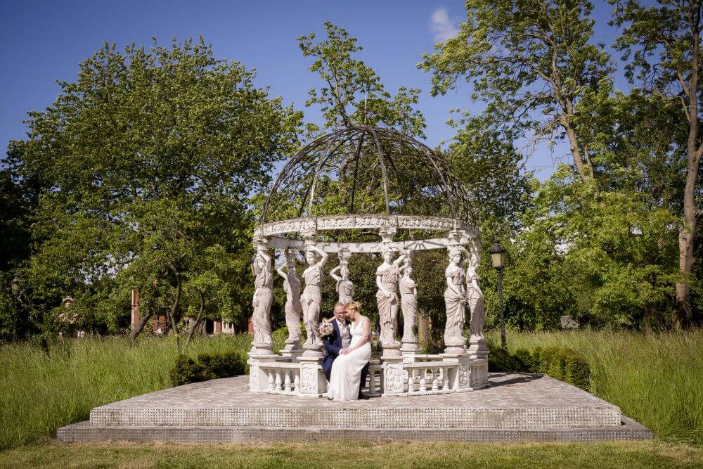 Bride and groom sitting under a marble gazebo-like structure with statues as columns. 