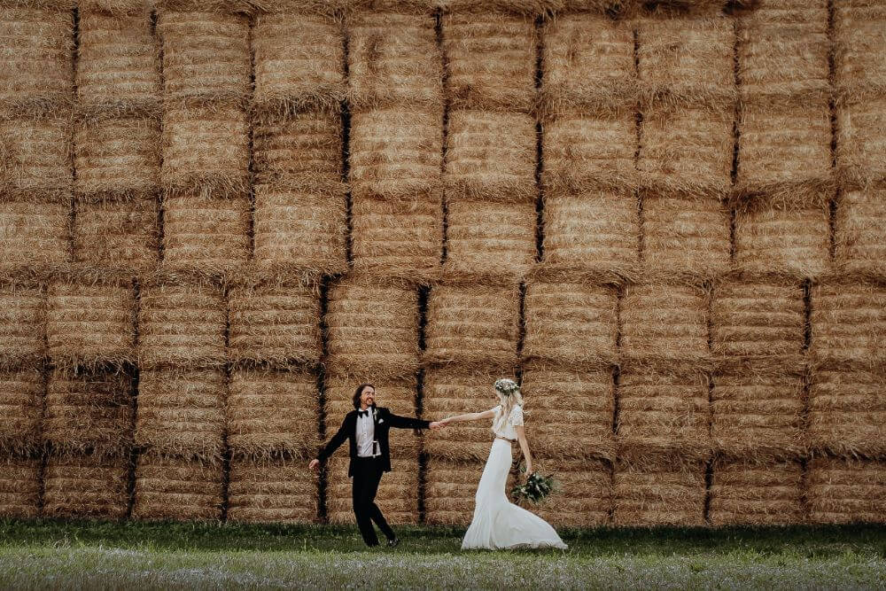 Bride and Groom hold hands. In the background, there are dozens of stacked piles of hay.