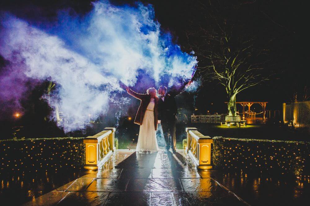 A newly wed bride and groom holding colourful smoke bombs in the air at night time.