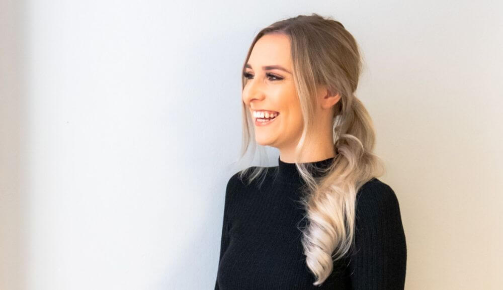 A left profile photo of a young blonde woman smiling against a white background. 