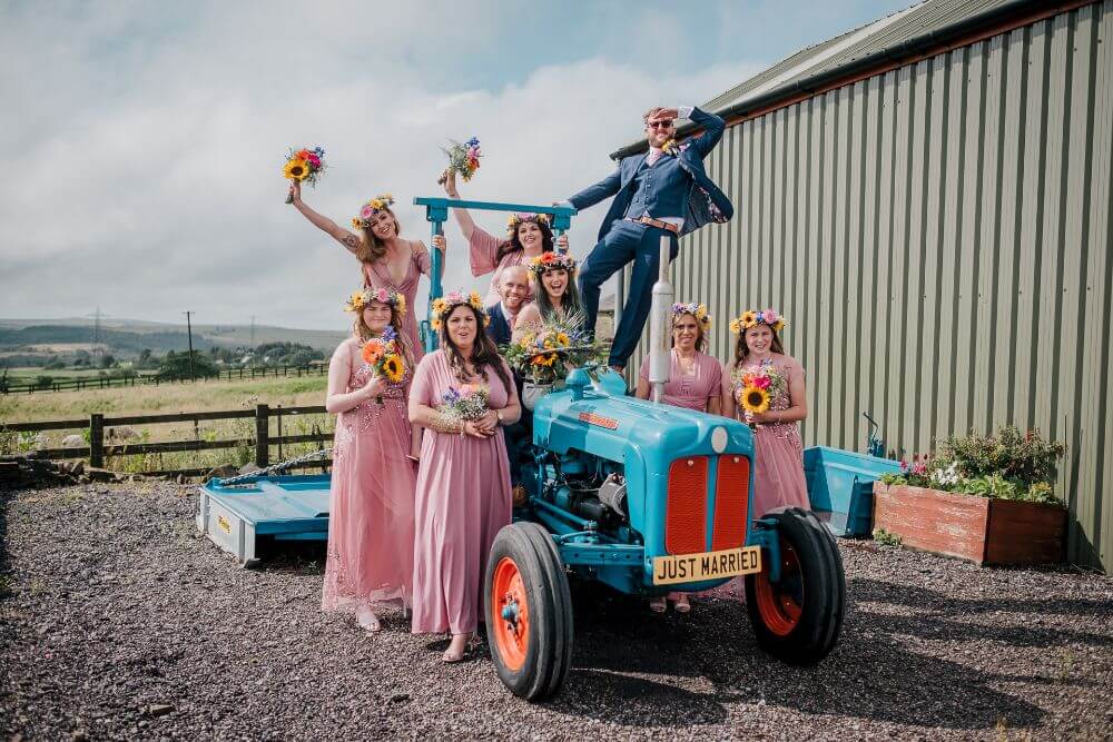 A wedding party (bride, groom, best man and bridesmaids) sat on a blue tractor with a 'Just Married' number plate. 