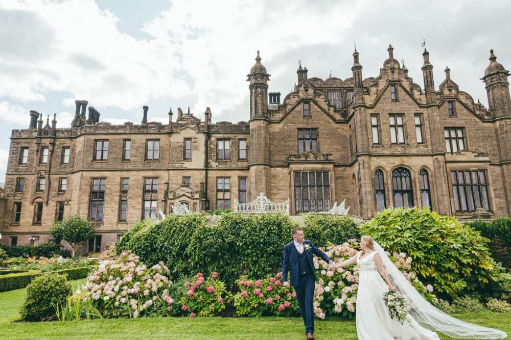 Bride and groom hold hands in a garden outside a castle. 