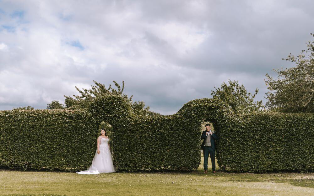 The bride and groom stand under a hedge outside.