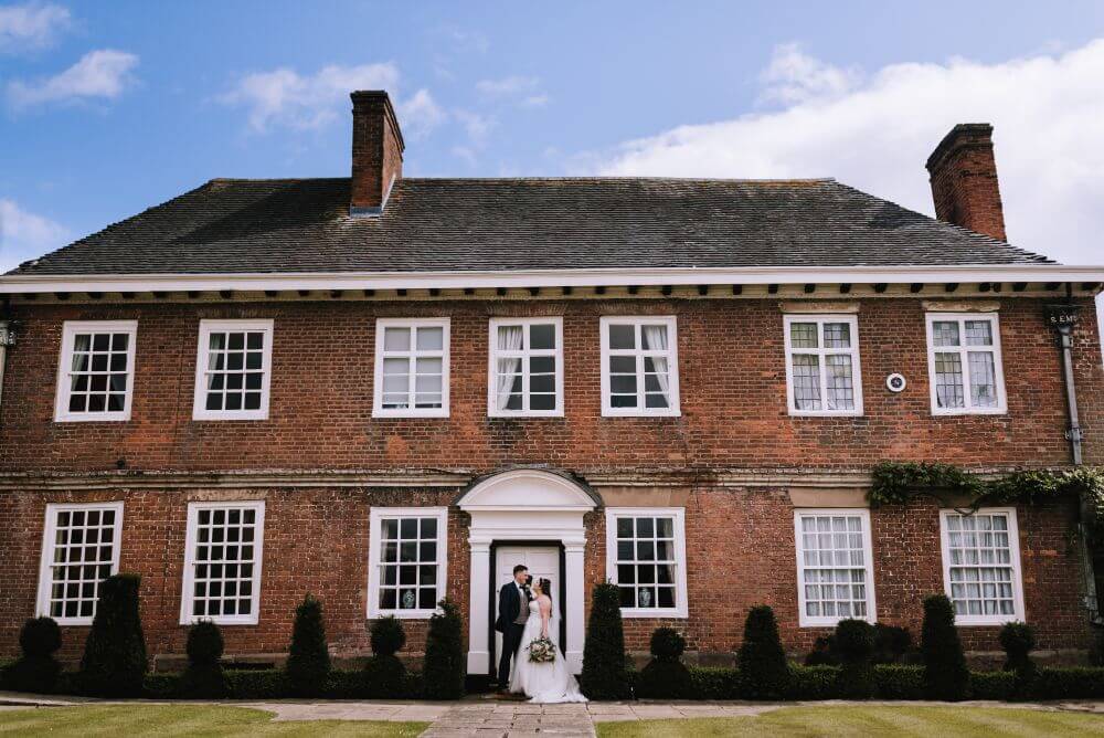 The bride and groom stand outside a big brick house with white windows.