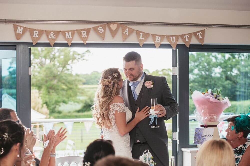 The bride and groom stand facing each other with the groom holding a glass of champagne.