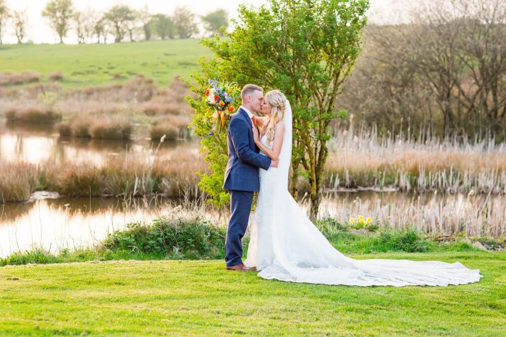 The couple kiss outside beside a lake.